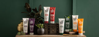 A lifestyle display of Grandpa Soap Co. hair care products, including Witch Hazel, Rosemary, Pine Tar, and Buttermilk shampoos and conditioners, arranged on a wooden cabinet with plants and vintage décor against a dark green wall.