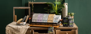 A wooden dresser holds stacked Grandpa Soap Co. soap bars in various colors, surrounded by potted plants, a woven cloth, and a mirror, all set against a green wall.