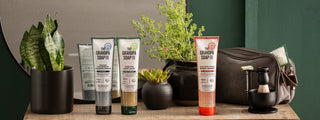 A wooden table displays Grandpa Soap Co. body washes, a black potted plant, small succulent, gray toiletry bag, and a shaving brush set, with a leafy plant and mirror in the background against a green wall.