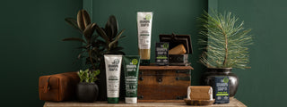 A lifestyle display of Grandpa Soap Co. Pine Tar body care products, including shampoo, conditioner, body wash, and bar soap, arranged on a rustic wooden cabinet with plants, a leather toiletry bag, and vintage wood accents against a dark green wall.