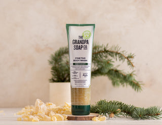 A tube of The Grandpa Soap Co. Pine Tar Body Wash stands upright on a surface with pine branches and resin pieces scattered around it, against a light, neutral background.