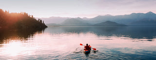 A person in a red kayak paddles across a calm lake at sunset, surrounded by mountains and trees with the sky reflected in the water.