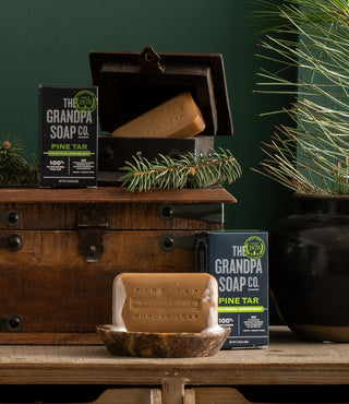 A bar of Grandpa Soap Co. Pine Tar soap sits in a brown dish on a wooden table, with two soap boxes and a wooden chest nearby. Pine branches decorate the rustic scene against a green background.