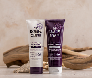Two tubes of The Grandpa Soap Co. Witch Hazel Clarify Shampoo and Conditioner sit on a table with driftwood.