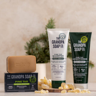 The Pine Tar Bar Soap and Haircare Bundle from The Grandpa Soap Co. is displayed on a light surface with a small pine tree and resin pieces in the background. The bundle includes bar soap, shampoo, and conditioner.
