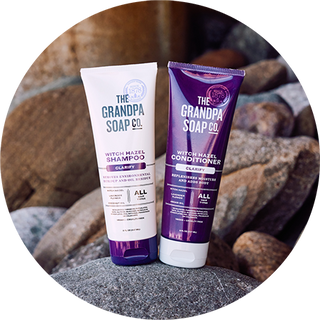 Two tubes of The Grandpa Soap Co. Witch Hazel Shampoo and Conditioner are placed upright on rocks. The shampoo tube is white and the conditioner tube is purple. Both are labeled Clarify and All Hair Types.
